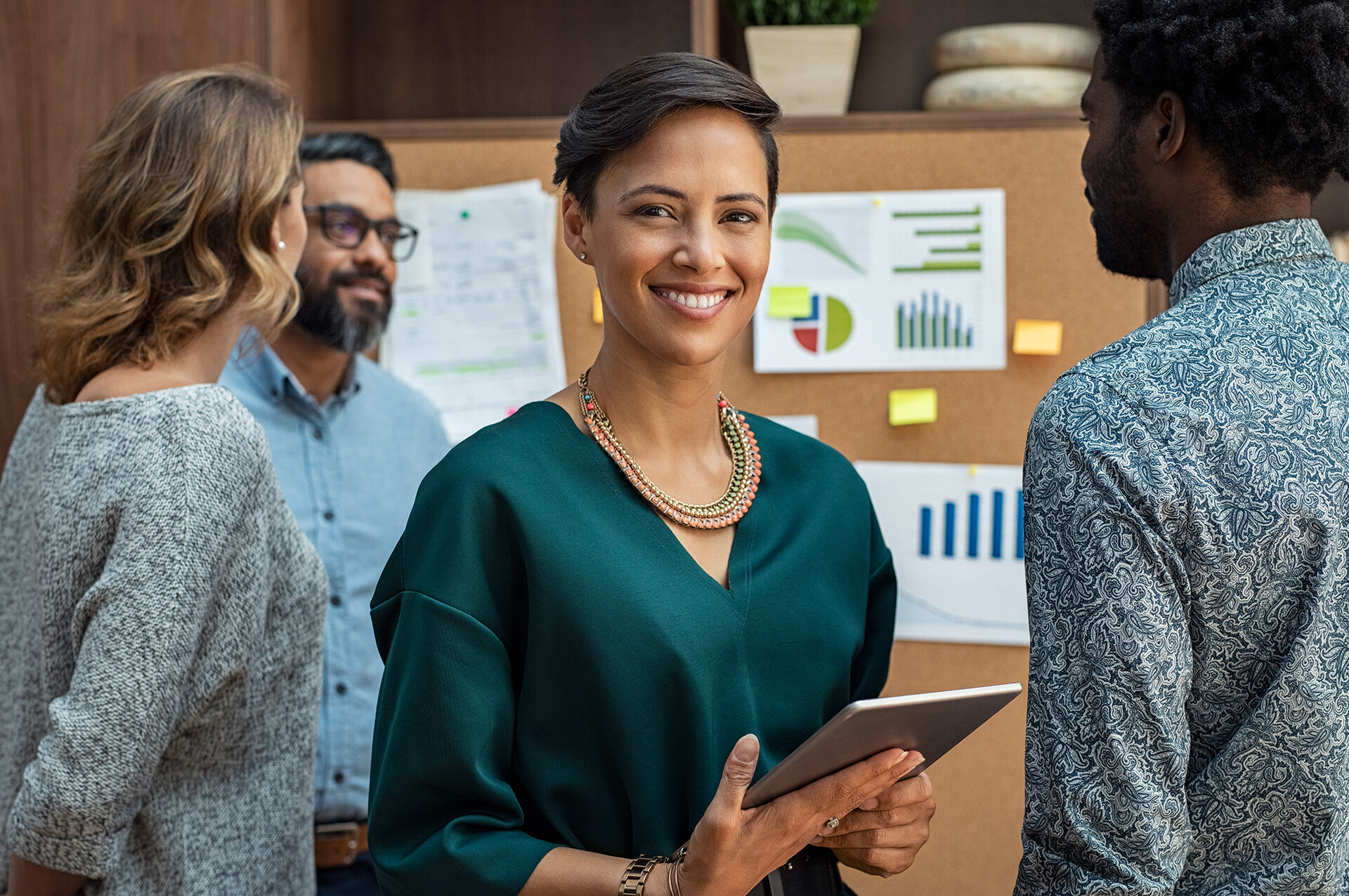 woman holding tablet beside coworkers standing in front of cork board with printed charts